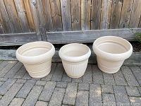 Three cream-colored plastic planters side by side on pavement, wood fence background.