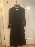 Full front view of the black long coat hanging on a white hanger on a door, showing overall length and fit.