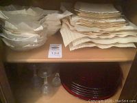 Shelf view showing stacked white and gold plates and bowls, red glass chargers, two wine glasses and decanter.