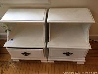 Photo of a pair of white wooden nightstands side by side against a wall, showing front view of both units with drawers and open shelves.
