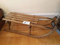 Full side view of wooden sled on hardwood floor showing slatted seat, runners and rope handle