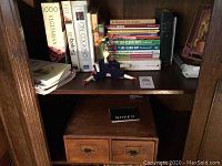 Vintage wooden card file recipe box with two drawers on lower shelf, assorted cookbooks on shelf above, small doll decoration on shelf.