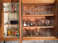 Wide view of cabinet with various glassware items spread on shelves including wine glasses, tumblers, and decorative pieces.