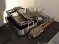 Stack of metal roasting pans, two clear glass bowls, and wooden handles or utensils wrapped in plastic.