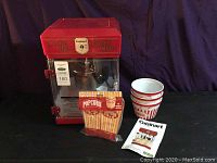 Front view of red Cuisinart Theatre Style popcorn maker with clear panels and metal kettle inside. Includes three striped popcorn cups, a package of popcorn bags, and user manual displayed alongside.