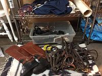 Photo shows pair of leather riding boots and horse tack pieces on a woven horse blanket under metal shelving.