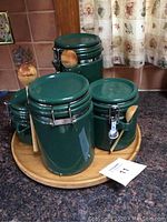 Four green ceramic canisters with metal latch closures and attached worn wooden spoons, arranged on wooden turntables. Largest turntable measures 18 inches in diameter.