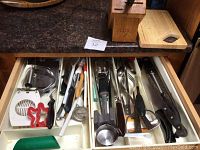 Photo of two kitchen drawers and wooden knife block showing various kitchen utensils including tongs, pastry blender, egg slicers, meat tenderizer, spoons, flatware, and other tools.