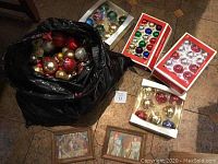 Photo showing black bag filled with assorted Christmas balls, three boxes of Christmas ornaments, and two framed Christmas artworks on floor