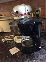 KitchenAid mixer with attached mixing bowl, metal whisk and white flat beater shown beside black Continental coffee maker on kitchen counter.