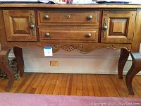 Front view of wooden side board showing two drawers, two cupboard doors, carved front apron, and curved legs on hardwood floor.