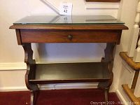 Two wood occasional tables with glass tops, single drawers, and bottom shelves photographed against a wall and staircase.