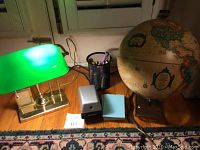 Wide view of entire lot showing green shade brass desk lamp, globe, pencil sharpener, note pads, pen holder, and extension cord on wood floor near window.