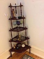 Corner wooden five-tier display shelf with assorted decorative items including wine glasses, crosses, clock, and figurines.