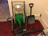 Photo showing Toro leaf blower on floor next to hose reel stand with green hose and snow shovel leaning against wall.