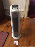 Front view of the Lasko white oscillating tower space heater standing on wooden floor with power cord visible.