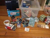 Photo showing the box contents with assorted light bulbs, batteries, and electrical tape arranged on floor near plastic container.