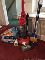 Full view of lot items: Dirt Devil vacuum, Charmin toilet paper, broom, ice scraper, and Lysol spray on carpeted floor near stairs.