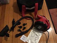 Photo of handheld red Scunci steamer, flexible hose, seven small black attachments, instruction manual, and part of the black carry bag with red handles.