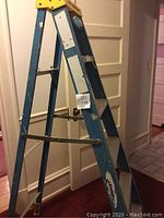 Photo showing side view of blue metal folding ladder with wear marks and Werner label on top step yellow cap.