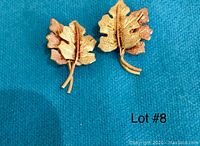 Front view of pair of 18 KT yellow and rose gold pierced earrings shaped as textured leaves with stem detail on blue fabric background.