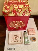 Holiday themed red box with gifts printed, and items laid in front: almond hand cream tube, tin of shea butter, pink soap box, and white ceramic soap dish.