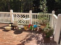 Outdoor deck photo displaying a collection of flower pots varying in size and color, a medium-sized metal table with lattice design, and multiple watering containers. A potted plant tree is also visible.