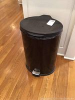 Metal black trash can closed with visible foot pedal on wooden floor.