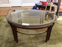 Full view of round coffee table with glass top, wooden legs, and metal support ring on beige carpeted floor.
