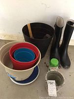 Multiple nested flower pots including plastic pots in beige, red, blue, black, and a single ceramic pot on a white saucer. Includes wooden-handled hand garden tools and a container of Osmocote fertilizer on the floor.