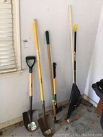 Photo showing all six tools lined up against wall in garage: Stanley long shovel, Ames short shovel, Ames rake, Rockford ax, Mr. Long Arm painter tool, Ames hoe.