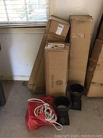 Three cardboard boxes containing boxed 4-foot pre-lit Christmas trees, two black resin urns on floor, red bag holding extension cords.