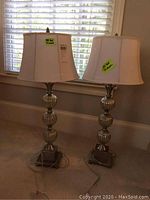 Pair of identical table lamps on carpeted floor in front of windows with white shades and metal/glass bases