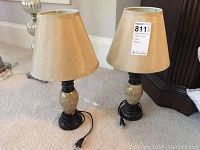 Pair of table lamps standing on beige carpet. Each has a beige lampshade, marble-like cream base with black accent and cord with plug. The lamps appear identical in style and size.