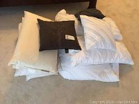 Stack of bedding items on carpet including white pillows, decorative brown pillow with tree design, and white comforter with sham.