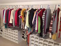 Row of assorted women's jackets, sweaters, cardigans, dresses, shirts mostly hanging on a rod with white cubby shelves below.