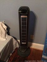 Holmes air purifier tower standing on the floor near a bed, dark gray with vent openings on the front