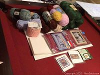 Wide view showing mixed skeins of yarn in multiple colors and several cross stitch kits with fabric and patterns on a red pool table surface.