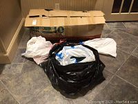 Large cardboard box and multiple plastic bags on tiled floor containing Christmas tree and decorations