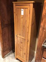 Front view of tall faux wood cabinet with vertical slat door and metal knob, positioned in a corner near other wooden furniture on tiled floor.