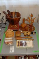 Overview of the collection showing the wood salad bowl with servers, wooden vase with carved floral pieces, a bottle, ceramic and wood sugar container, salt and pepper shakers, knives in box, napkin holder, and marble and wood coasters arrayed on a grey table surface.