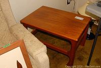 Side angle view of a teak Midcentury Modern side table beside a couch, showing the wood grain and rounded edging on top.