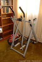 Side view of the stepper machine showing metal frame and foot pedals near a wooden bookshelf.