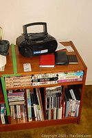 Shelf unit containing black Sony portable stereo on top, assortment of DVDs, CDs, VHS tapes, and LP vinyl records below, plus two notebooks on top near stereo.