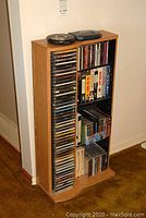 Vertical wood-look stand holding over 50 CDs and several VHS tapes, placed against a wall with carpeted floor visible.