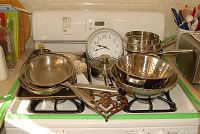 Full view of the collection arranged on a gas stove, showing stainless steel pots, pans, colanders, and a wall clock placed at the back.