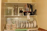 Kitchen cabinet shelf with plastic, glass measuring cups, ceramic mugs, and Corningware dishes with covers.