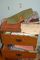 Stack of various tablecloths and cloths on a wooden dresser with drawers partly open, showing multiple folded textile items.