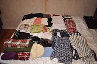 Photo shows an array of folded women's clothing in medium size including various tops, leggings, scarves, hats, gloves, packaged underwear, and handbags laid out on a bed.