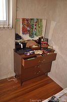 Wooden 3 drawer dresser with open top drawer and assorted items on top, mirror attached behind.